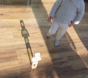 Colour Block Image: A child in light-coloured clothes stands on a wooden floor next to a small stack of coloured blocks casting a shadow in the sunlight.