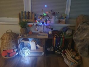 An indoor area set up on a low windowsill and against a wall. Resources including wooden baskets and blocks, books natural loose parts are displayed, illuminated by a multi-coloured light tree